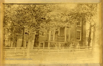 'This was the last female seminary in Morgantown. It was owned and operated by Mrs. E.L. Moore, mother of Susan Maxwell Moore. Mrs. Moore had been the principal of Woodburn Female Seminary located where Woodburn Hall now stands. This last seminary was on the corner of High and Foundry Streets, facing High Street. It burned in April 1889. Back row left to right are: Mrs. Sarah Moore (grandmother), Mrs. Elizabeth Deven McGee, Mrs. E.L. Moore, unidentified, Mrs. M. J. Brand, Willa Brand, George M. Brand, and Lieutenant Will Moore of the Navy. Front row left to row are: Mrs. Will Moore (Lois), Willie Moore, Elizabeth Thomas, Mrs. M. Sweeney, and Susan Maxwell Moore.'