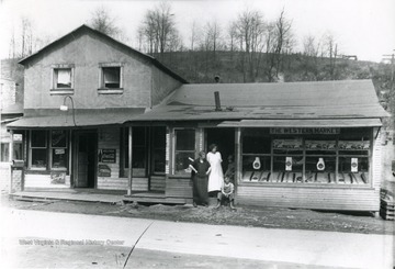 Exterior view of the Moss Restaurant to the left and the Western Market on the right. Two women and two children in the doorway of the Western Market.