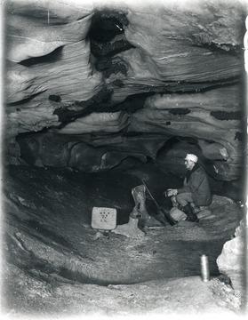 Man in a cave with a gun, satchel, and light.