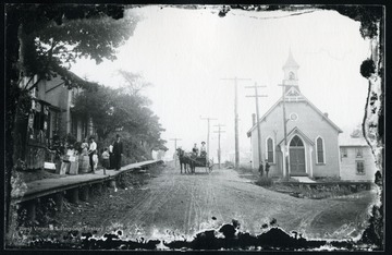A group of people are seen on a raised wooden sidewalk across from the Methodist Episcopal Church.