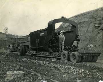 Man stands on the back of the McCoy Brothers Truck that is transporting a large Bucyrus Erie shovel.