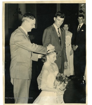 Women kneeling down, while a man places a crown on her head.