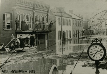 A flood on Main Street in Wellsburg, W. Va. People in row boats and on second story porch at left.