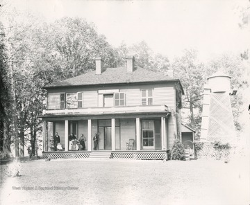 Portrait of a family on the porch of a home in Greenbrier County. An old man stands beside the house to the left, and a pet dog lies in the front yard.