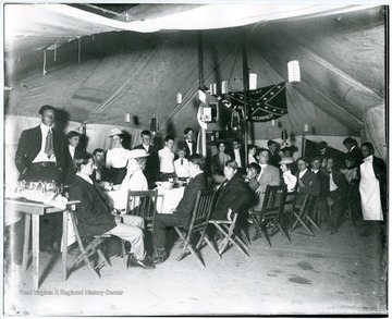 People gathered for a party under a Confederate flag bearing the inscription "Camp Greenbrier" with two African-Americans dressed as servers standing in the background.
