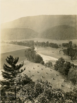 View of harvested fields and a tree-lined river running through the center of this scene in Hampshire County. Tree-covered mountains are seen in the distance.