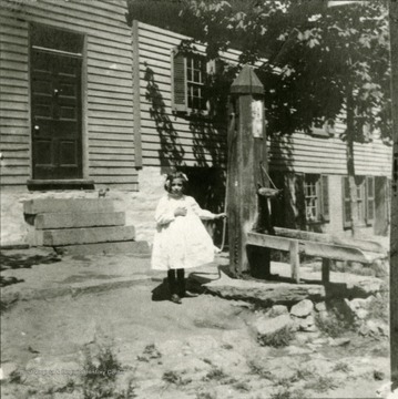 Young girl, Rachel Snyder, stands by Grant's pump. The Grant house, later burned, is seen in the background. The house was located on the North side of German Street, between Duke and Church Streets.