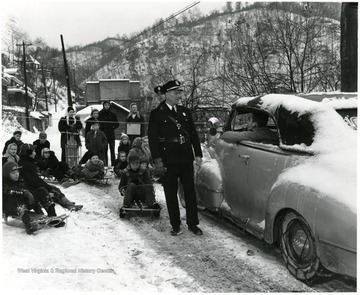 Children on sleds look on as police officers talk to a man in his car.