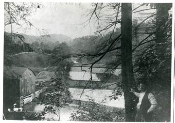 A view of the Barrackville Covered Bridge in Marion County, West Virginia. An unidentified man is leaning on a tree above the covered bridge. Mountains are in the background.