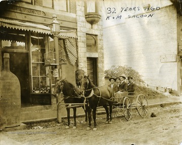 'A picture of surrey and high-stepping horses owned by James H. Moyer;picture made in front of Kelley and Moyer bar, in 1900, before prohibition days. Note large demijohn in front of door; bird cages house a parrot in each one. The building is located at 400 Bland Street, must below the Daily Telegraph Printing Company, and in the same block with the Bland Street Methodist Church. Ash M. Prince, pioneer resident of the city, erected the building. After prohibition days, the building was used by the Hawkins Undertaking Company until they built their mortuary at the corner of Bland and Preston Streets, and the building as it now stands is owned by the Sweet Shop. In the picture are; front seat, left to right-James H. Moyer and P. J. Kelley, owners of the surrey and the bar; rear seat, left to right, M. B. Postlethwaite, city sergeant, and Robert Dunn, chief of police.'