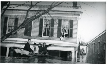 Four men in a boat near a house where two women await rescue.