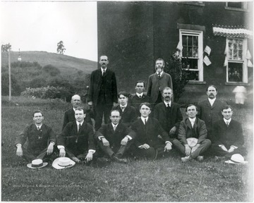 First Row, Left to Right:  O. T. Sines (day guard), George Travis (eng.), H. O. Vantramp (eng.), Frank Moore (baker), unknown (teacher), W. R. Collins (guard).  Second Row, Left to Right:  Mr. Walter (stable boss), Ralph Kunst (guard), C. E. Bunner (chief eng.), H. L. McMillin (boy's cook).  Third Row, Left to Right:  Mr. Deering (guard), E. Whitescarver (teacher), Mr. Sheppard (shoe shop supervisor).'  The men pose for a group portrait as the boys look out the windows of the school behind them. 