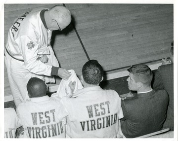 West Virginia University Basketball Trainer Whitey Gwynne is handing out towels to players sitting on the bench. 'Taylor Publishing Company, Job Number 07206, Picture Number 2, page 209.'