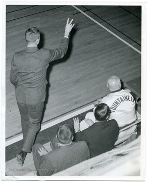 West Virginia University Basketball Coach Bucky Waters is signaling plays to his players, while Assistant Coach Sonny Moran, an unidentified coach, and Whitey Gwynne, the trainer looks on from the bench.