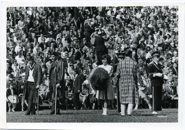 Crowd looks on during a football game. Fi Batar Cappar men in long printed coats holding canes stand near a cheerleader. 'Taylor Publishing Company 07206, Picture Number and Page Number are omitted.'