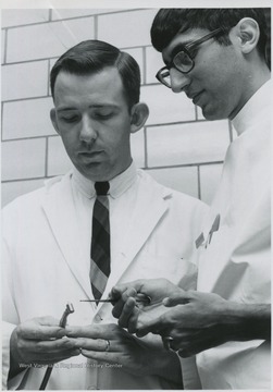 'Progress in Dentistry- Contrasting ultra- speed bur, or drill, used regularly in dental clinics at WVU Medical Center with manual type used by dentists in the 1800s are Dr. Clarence R. McCurdy Jr. (left) instructor in operative dentistry, and Anthony Alleruzzo of Erie, Pa., third- year dental student.  The old instrument, with its special cupped ring that slipped over the middle finger and protected the palm of the hand, is part of a collection given to the WVU School of Dentistry by descendants of Dr. John Matthew Gay Fairfax, once a dentist in Reedsville, Preston County, W. Va.'