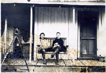 Two men seated in a porch swing.  Survey equipment nearby.