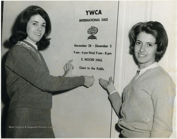 Two unidentified women gesturing to the YWCA poster, announcing an upcoming sale in E. Moore Hall.