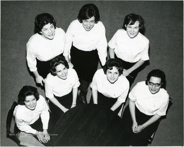 Members of Associated Women Students Executive Council meet around a round table dressing alike with uniform white top and dark bottom.  They are looking up and smiling to the camera.  