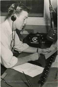 A student with a headphone sitting at a broadcast board reads an article in his hand.