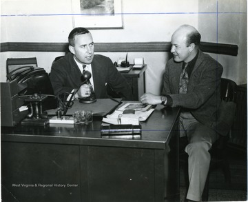 Two WVU faculty members sit at a desk while one on the left Joseph Gluck holds a microphone.