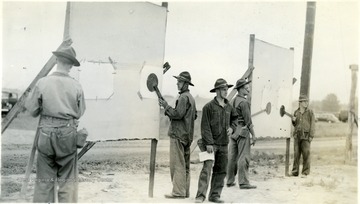 Cadets in a field appeared to be preparing target for shooting training; piece of papers are taped on the boards; some cadets place disc over the paper. 