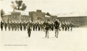 Cadets with a gun on shoulder march on the Drill Field. There are three saluting cadets leading the rest.