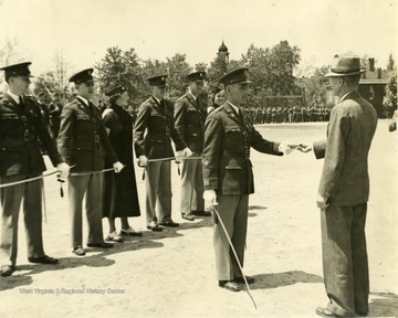 Cadet Captain Nels at center holding sword and passing object to a civilian man.