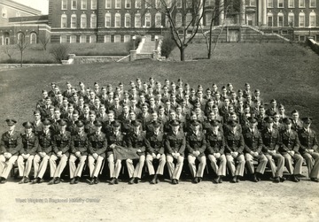 The cadets of company H sit on bleachers with officers for a group portrait on the Drill Field with Stalnaker Hall in the background. See original for identification.