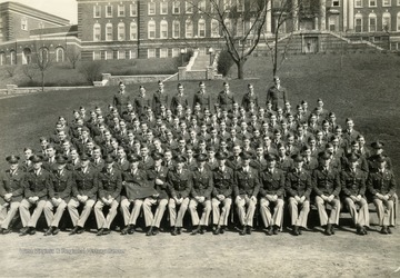 The cadets of company C sit on bleachers with officers for a group portrait on the Drill Field with Stalnaker Hall in the background. See original for identification.