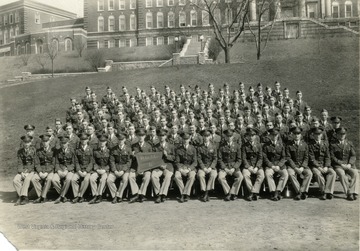 The cadets of company B sit on bleachers with officers for a group portrait on the Drill Field with Stalnaker Hall in the background. See original for identification.