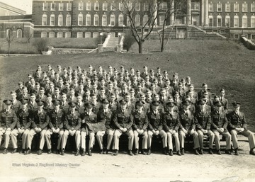 The cadets of company G sit on bleachers with officers for a group portrait on the Drill Field with Stalnaker Hall in the background. 