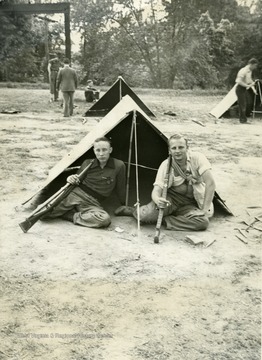 Two cadets with a gun on their shoulders sit in front of a pitched tent.