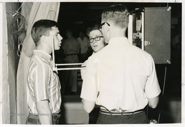 A student stands facing a camera for his ID picture while two photo technicians adjust.