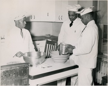 Three students stand around a table sifting flour over a bowl.