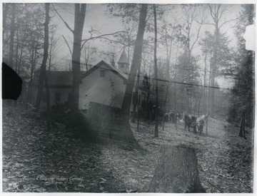 A school building in the woods; there are students in front of the building and a few horse drawn carriages.