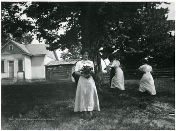 Miss Kennedy from Fairmont stands in a field with bouquet of flowers in hands while others engage in some activities in the background.