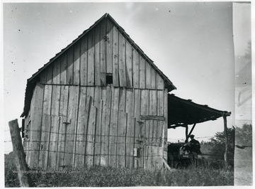 A view of barn; a man and a farm machine are under an awning on front.