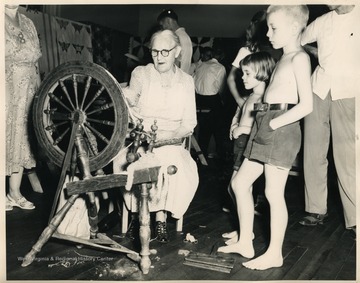 'Youngsters catch a glimpse of a vanished art. The spinning wheel hums busily before a background of hand-made quilts, representing a variety of designs once familiar to the folk.'