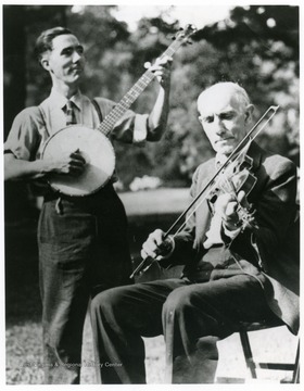 Edden Hammons seated playing fiddle with his son James standing playing banjo.