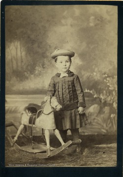 A cabinet card portrait of an unidentified little boy dressed in velvet trimmed dress and straw hat, standing with a rocking horse.
