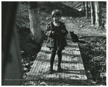 A little girl standing on a walkway in the woods with a black cat in the background.