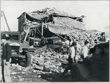 A photograph of a destroyed building with an automobile among the wreckage and men walking about.