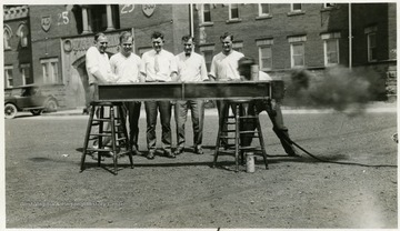A group of men involved in testing a piece of unidentified equipment. WVU Armory visible in background.