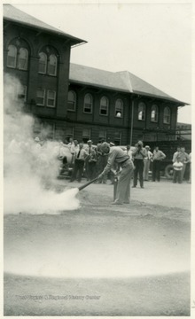 A photograph of a man putting out a fire with an extinguisher as part of fire safety training with a large group observing in front of WVU Mechanical Hall.