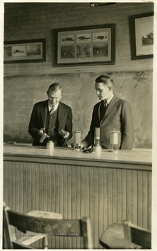 A photograph of two men standing in front of a classroom working on a safety lamp.