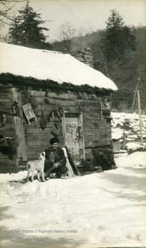 A photograph of Dana Hicks, holding a hunting rifle, with his dog outside a cabin.