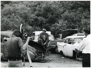 A photograph of A. James Manchin being filmed among a group of scrapped automobiles.
