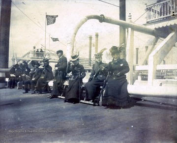 A photograph of unidentified men and women seated on the deck of a ship.  The smoke stacks of a steam boat can be seen in the background.