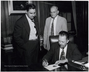 A photograph of Governor Patteson (seated) signing a piece of paper, with Eugene Carter (left), President of the West Virginia Federation of Labor, and another man standing behind him. 